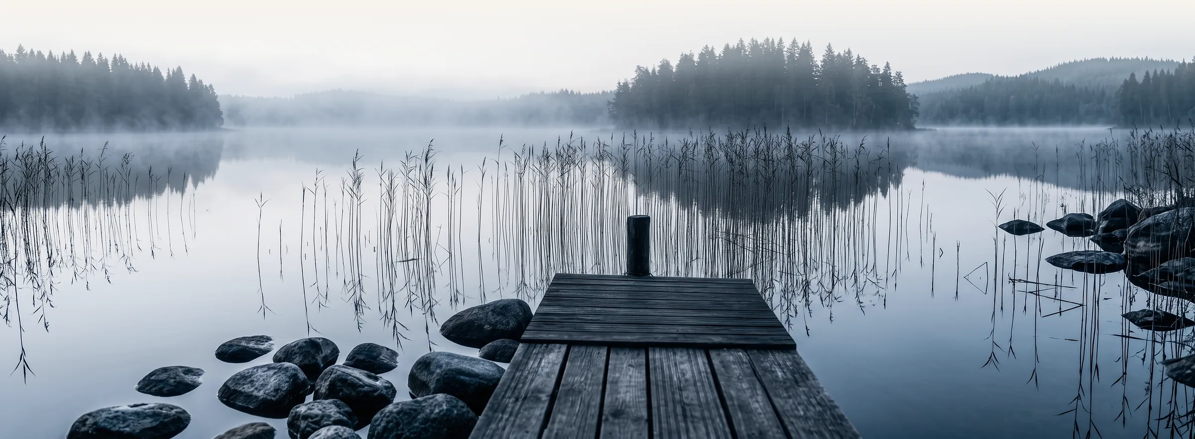 Sumuinen järvi aamuhämärässä: puinen laituri, kaislikko, rantakiviä ja metsän siluetti veden takana.